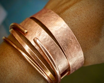 Peut inclure: Un ensemble de quatre bracelets en cuivre avec une finition martelée. Les bracelets sont empilés les uns sur les autres et sont portés au poignet.