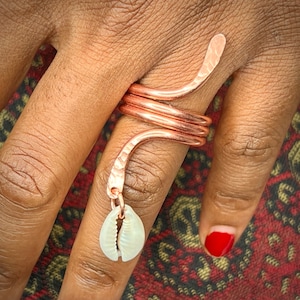 May include: A copper ring with a hammered finish and a white shell charm hanging from it. The ring is worn on a finger.