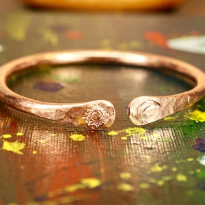 May include: A hammered copper bangle with a sun and moon design. The bangle is open and has a textured finish.
