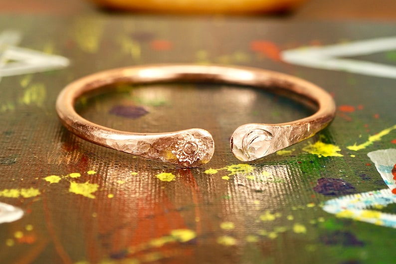 May include: A hammered copper bangle with a sun and moon design. The bangle is open and has a textured finish.