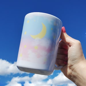 May include: A white ceramic mug with a pastel pink, blue, and yellow watercolor design of clouds and a crescent moon. The mug has a handle and is held in a hand against a blue sky with white clouds.