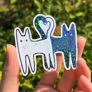 Könnte beinhalten: Ein gestanzter Aufkleber mit zwei stilisierten Katzen, die nebeneinander stehen. Eine Katze ist weiß mit regenbogenfarbenen Glitzerakzenten, die andere schwarz mit blauem und grünem Glitzer. Ihre Schwänze bilden ein Herz über ihren Körpern.
