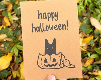 Puede incluir: Una tarjeta de felicitación rectangular de color tostado con el texto impreso en negro happy halloween! sobre un dibujo lineal negro de un gato sentado dentro de una calabaza tallada con una tela drapeada en el lateral.