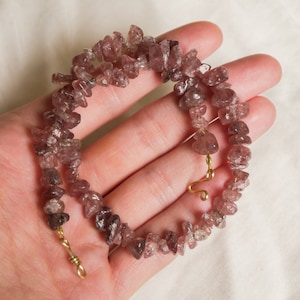 Può includere: Una collana realizzata con piccole pietre di quarzo irregolari, rosa chiaro e trasparenti. Le pietre sono infilate su un filo d'oro e hanno una chiusura dorata.
