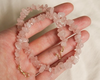 May include: A delicate pink rose quartz chip necklace with a gold clasp. The necklace is made of natural stones and is strung on a thin wire.