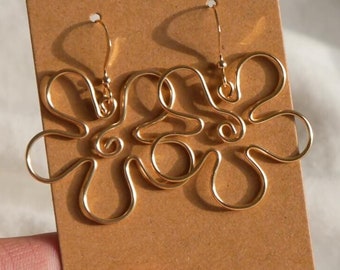 Puede incluir: Pendientes de alambre dorados con diseño floral. Los pendientes se muestran en una tarjeta marrón con un agujero en la parte superior. El fondo es una tela blanca texturizada.