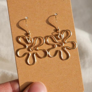 Auf einem weißen Leintuch hält eine Hand eine Karton Displaykarte mit goldenen Ohrringen. Die Ohrringe haben Anhänger aus Draht in Form einer Blume mit runden Blütenblättern.