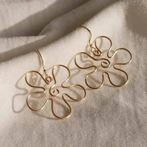 Auf einem weißen Leintuch liegen ein Paar goldene Ohrringe. Die Ohrringe haben Anhänger aus Draht in Form einer Blume mit runden Blütenblättern.