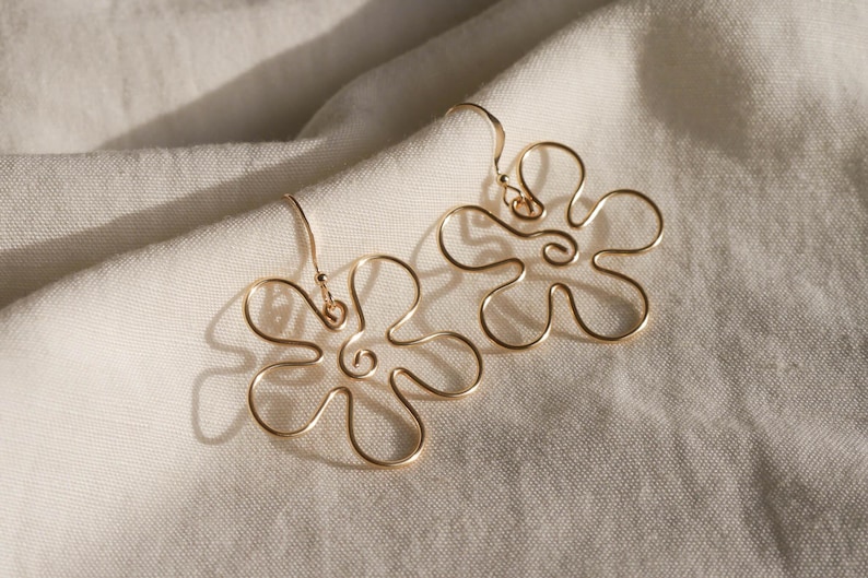 Auf einem weißen Leintuch liegen ein Paar goldene Ohrringe. Die Ohrringe haben Anhänger aus Draht in Form einer Blume mit runden Blütenblättern.