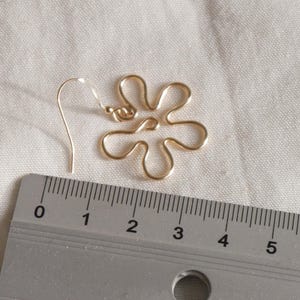 Auf einem weißen Leintuch liegt ein goldener Ohrring. Der Anhänger des Ohrrings ist aus Draht in Form einer Blume mit runden Blütenblättern. Daneben liegt ein Lineal. Man kann ablesen, dass der Ohrring circa 4 Zentimeter lang ist.