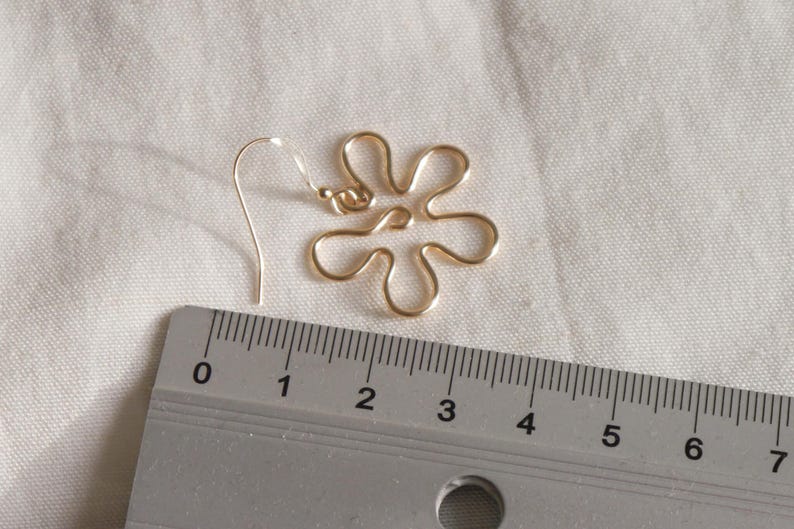 Auf einem weißen Leintuch liegt ein goldener Ohrring. Der Anhänger des Ohrrings ist aus Draht in Form einer Blume mit runden Blütenblättern. Daneben liegt ein Lineal. Man kann ablesen, dass der Ohrring circa 4 Zentimeter lang ist.