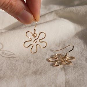 Auf einem weißen Leintuch liegt ein goldener Ohrring. Zwei Finger halten den zweiten Ohrring. Die Ohrringe haben Anhänger aus Draht in Form einer Blume mit runden Blütenblättern.
