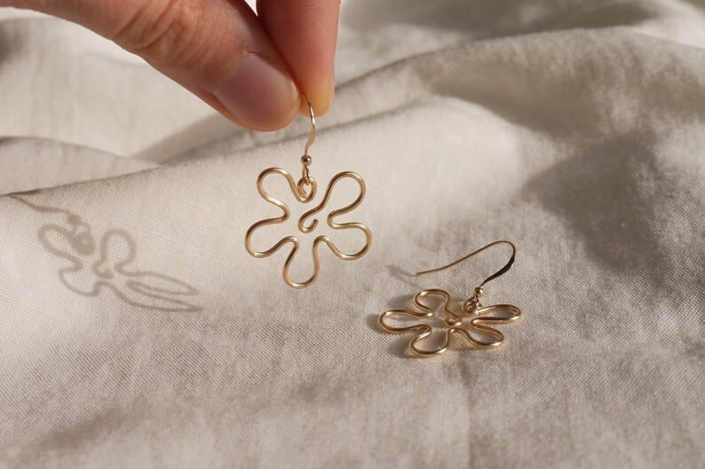 Auf einem weißen Leintuch liegt ein goldener Ohrring. Zwei Finger halten den zweiten Ohrring. Die Ohrringe haben Anhänger aus Draht in Form einer Blume mit runden Blütenblättern.