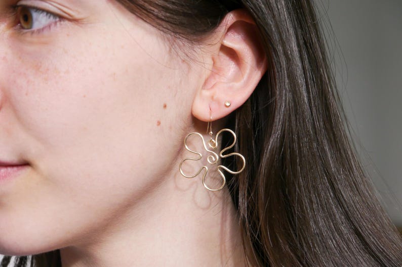Eine junge Frau trägt einen goldenen Ohrring. Der Anhänger des Ohrrings ist aus Draht in Form einer Blume mit runden Blütenblättern.