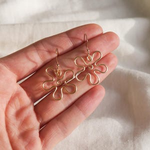 Auf einem weißen Leintuch hält eine Hand ein Paar goldene Ohrringe. Die Ohrringe haben Anhänger aus Draht in Form einer Blume mit runden Blütenblättern.