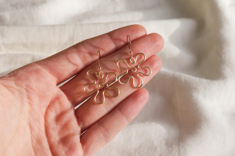 Auf einem weißen Leintuch hält eine Hand ein Paar goldene Ohrringe. Die Ohrringe haben Anhänger aus Draht in Form einer Blume mit runden Blütenblättern.