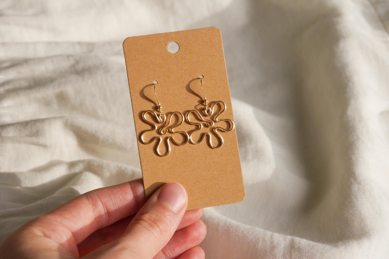 Auf einem weißen Leintuch hält eine Hand eine Karton Displaykarte mit goldenen Ohrringen. Die Ohrringe haben Anhänger aus Draht in Form einer Blume mit runden Blütenblättern.