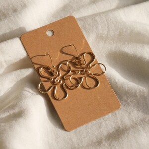 Auf einem weißen Leintuch liegt eine Karton Displaykarte mit goldenen Ohrringen. Die Ohrringe haben Anhänger aus Draht in Form einer Blume mit runden Blütenblättern.