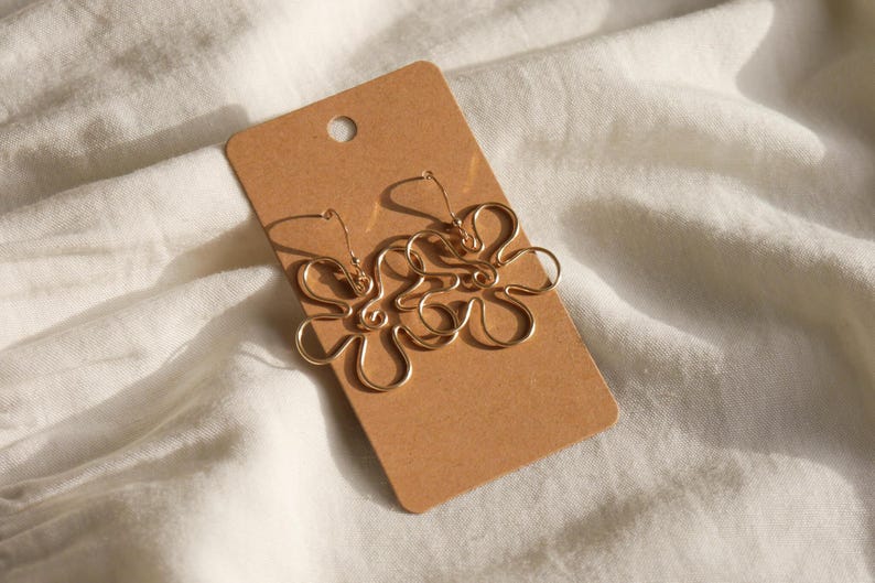 Auf einem weißen Leintuch liegt eine Karton Displaykarte mit goldenen Ohrringen. Die Ohrringe haben Anhänger aus Draht in Form einer Blume mit runden Blütenblättern.