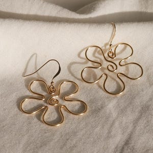 Auf einem weißen Leintuch liegen ein Paar goldene Ohrringe. Die Ohrringe haben Anhänger aus Draht in Form einer Blume mit runden Blütenblättern.