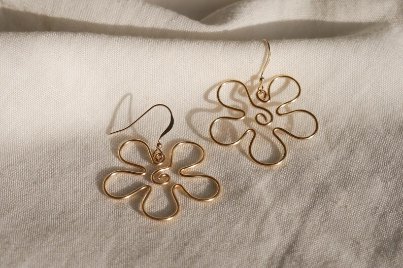 Auf einem weißen Leintuch liegen ein Paar goldene Ohrringe. Die Ohrringe haben Anhänger aus Draht in Form einer Blume mit runden Blütenblättern.