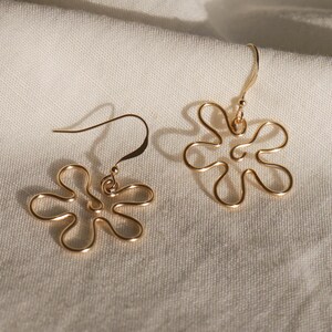 Auf einem weißen Leintuch liegen ein Paar goldene Ohrringe. Die Ohrringe haben Anhänger aus Draht in Form einer Blume mit runden Blütenblättern.