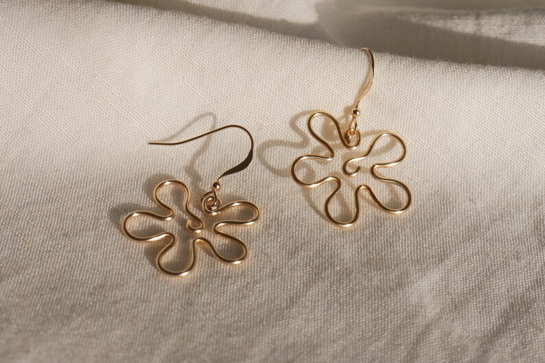 Auf einem weißen Leintuch liegen ein Paar goldene Ohrringe. Die Ohrringe haben Anhänger aus Draht in Form einer Blume mit runden Blütenblättern.