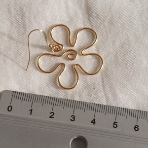 Auf einem weißen Leintuch liegt ein goldener Ohrring. Der Anhänger des Ohrrings ist aus Draht in Form einer Blume mit runden Blütenblättern. Daneben liegt ein Lineal. Man kann ablesen, dass der Ohrring circa 4 Zentimeter lang ist.