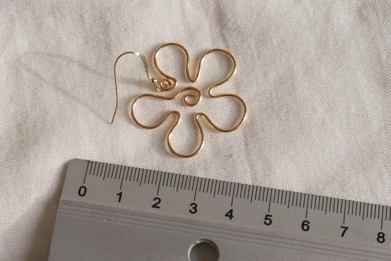 Auf einem weißen Leintuch liegt ein goldener Ohrring. Der Anhänger des Ohrrings ist aus Draht in Form einer Blume mit runden Blütenblättern. Daneben liegt ein Lineal. Man kann ablesen, dass der Ohrring circa 4 Zentimeter lang ist.