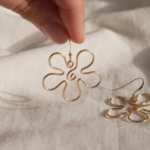 Auf einem weißen Leintuch liegt ein goldener Ohrring. Zwei Finger halten den zweiten Ohrring. Die Ohrringe haben Anhänger aus Draht in Form einer Blume mit runden Blütenblättern.