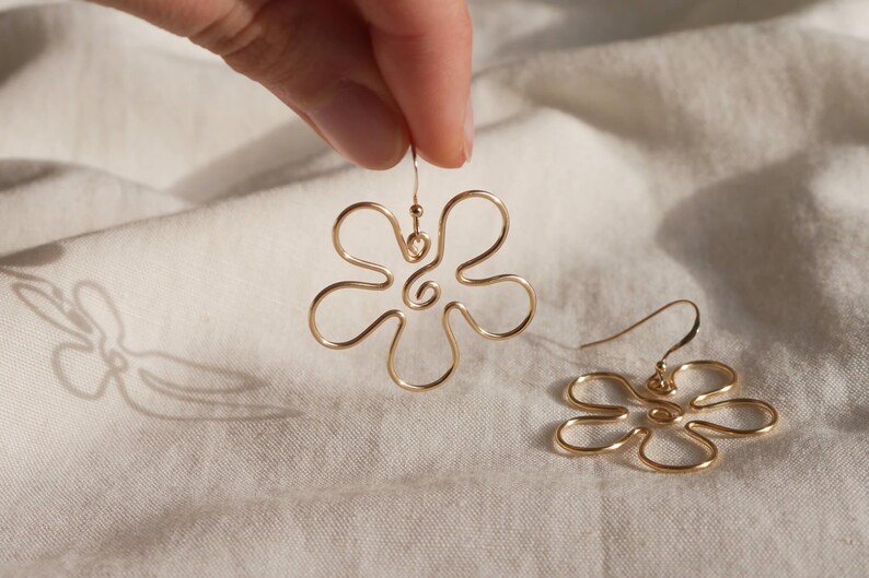 Auf einem weißen Leintuch liegt ein goldener Ohrring. Zwei Finger halten den zweiten Ohrring. Die Ohrringe haben Anhänger aus Draht in Form einer Blume mit runden Blütenblättern.