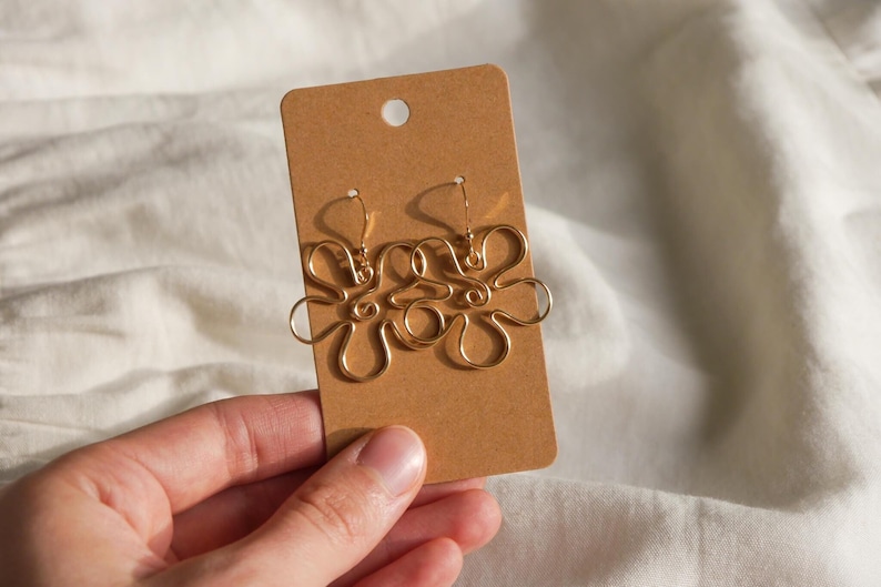 Auf einem weißen Leintuch hält eine Hand eine Karton Displaykarte mit goldenen Ohrringen. Die Ohrringe haben Anhänger aus Draht in Form einer Blume mit runden Blütenblättern.