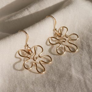 Auf einem weißen Leintuch liegen ein Paar goldene Ohrringe. Die Ohrringe haben Anhänger aus Draht in Form einer Blume mit runden Blütenblättern.