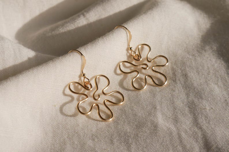 Auf einem weißen Leintuch liegen ein Paar goldene Ohrringe. Die Ohrringe haben Anhänger aus Draht in Form einer Blume mit runden Blütenblättern.