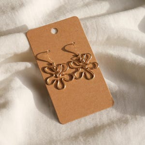 Auf einem weißen Leintuch liegt eine Karton Displaykarte mit goldenen Ohrringen. Die Ohrringe haben Anhänger aus Draht in Form einer Blume mit runden Blütenblättern.