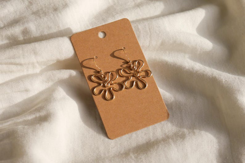 Auf einem weißen Leintuch liegt eine Karton Displaykarte mit goldenen Ohrringen. Die Ohrringe haben Anhänger aus Draht in Form einer Blume mit runden Blütenblättern.