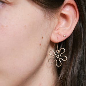 Eine junge Frau trägt einen goldenen Ohrring. Der Anhänger des Ohrrings ist aus Draht in Form einer Blume mit runden Blütenblättern.