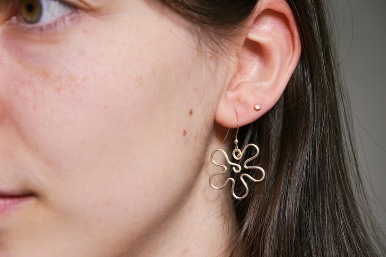 Eine junge Frau trägt einen goldenen Ohrring. Der Anhänger des Ohrrings ist aus Draht in Form einer Blume mit runden Blütenblättern.