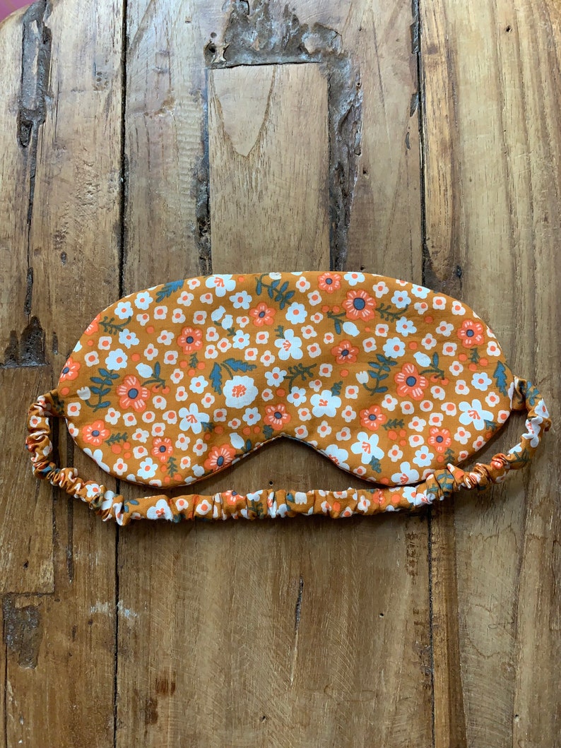 May include: An eye mask with a floral pattern in shades of orange, white, and blue. The mask has an elastic band for a comfortable fit. The sleep mask is designed to block out light for better sleep.