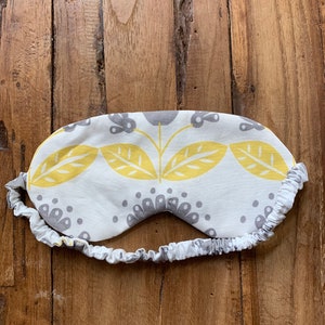 May include: A white sleep mask with a yellow and gray floral pattern. The mask has an elastic band and is designed to block out light. The sleep mask is made of fabric and is designed for comfort.