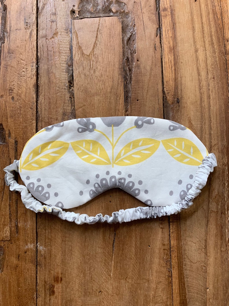 May include: A white sleep mask with a yellow and gray floral pattern. The mask has an elastic band and is designed to block out light. The sleep mask is made of fabric and is designed for comfort.