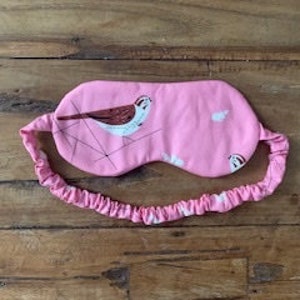 May include: Pink sleep mask with a bird illustration and matching elastic band. The mask features a white and brown bird design on a pink background. The elastic band is also pink.