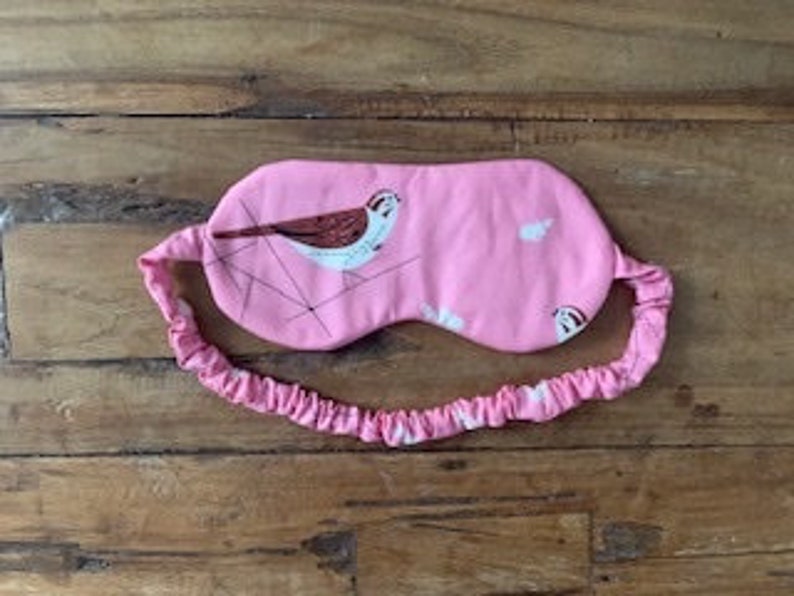 May include: Pink sleep mask with a bird illustration and matching elastic band. The mask features a white and brown bird design on a pink background. The elastic band is also pink.