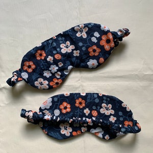 May include: Two navy blue sleep masks with a floral pattern of white and orange flowers. The masks have an elastic band and are designed to block out light. The fabric has a delicate, soft appearance.