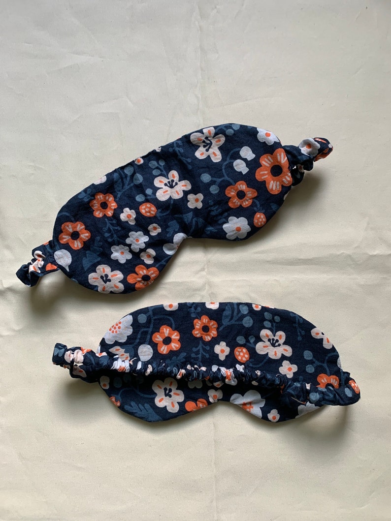 May include: Two navy blue sleep masks with a floral pattern of white and orange flowers. The masks have an elastic band and are designed to block out light. The fabric has a delicate, soft appearance.