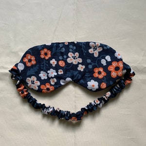 May include: A navy blue sleep mask with a floral pattern in white and orange. The mask has a matching elastic band for a comfortable fit. The sleep mask is designed to block out light for better sleep.