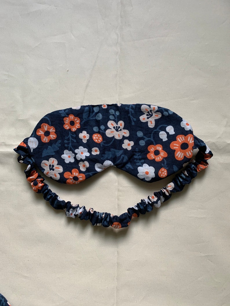 May include: A navy blue sleep mask with a floral pattern in white and orange. The mask has a matching elastic band for a comfortable fit. The sleep mask is designed to block out light for better sleep.