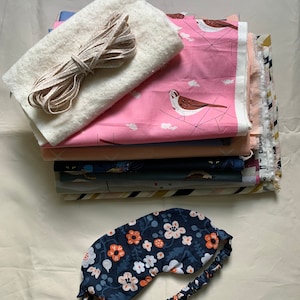 May include: A stack of fabric pieces in various colors and patterns, including a pink fabric with bird illustrations. A white, fluffy fabric and a length of beige ribbon are also present. A navy blue floral sleep mask with an elastic band rests at the bottom.