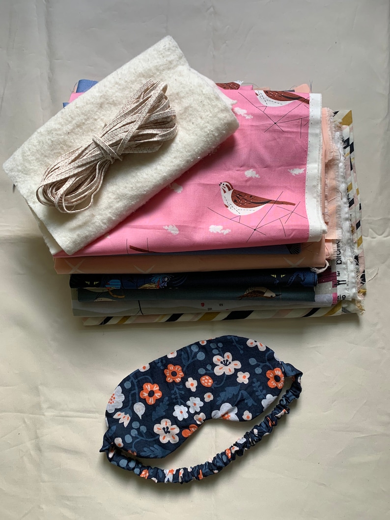 May include: A stack of fabric pieces in various colors and patterns, including a pink fabric with bird illustrations. A white, fluffy fabric and a length of beige ribbon are also present. A navy blue floral sleep mask with an elastic band rests at the bottom.