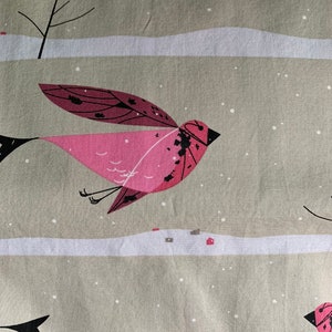 May include: Fabric with a repeating pattern of stylized pink birds in flight against a winter landscape. The birds have dark accents and are set against a neutral background with white snow and small houses.